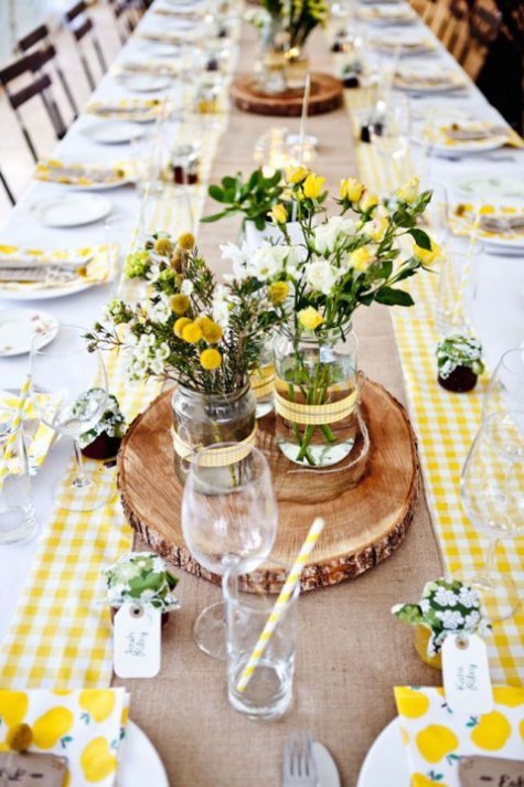 a checked yellow table runner with a burlap one on top, wood slices and yellow roses and billy balls