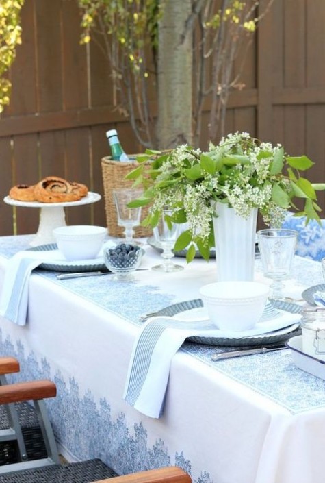 a white and blue printed tablecloth and napkins, a greenery and flower centerpiece in a glass vase