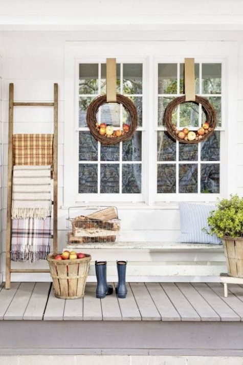 a cozy porch with vine and pumpkin wreaths_ a wooden basket with apples_ plaid blankets and firewood in a mtel crate